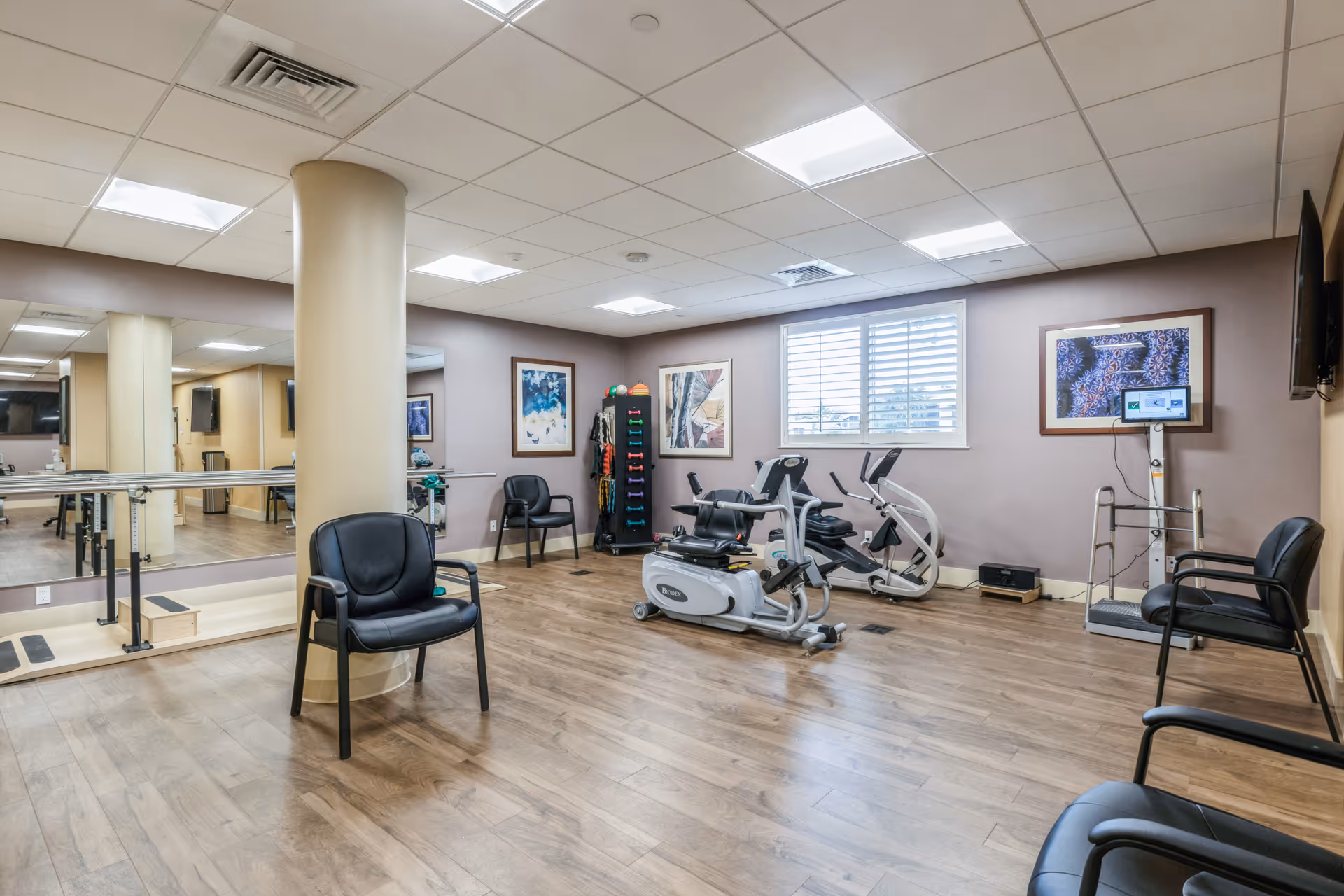 A well-lit exercise room with wooden flooring, featuring stationary exercise bikes, a rack of colorful dumbbells, several black chairs, a large mirror on one wall, and framed artwork. A window with white blinds lets in natural light.