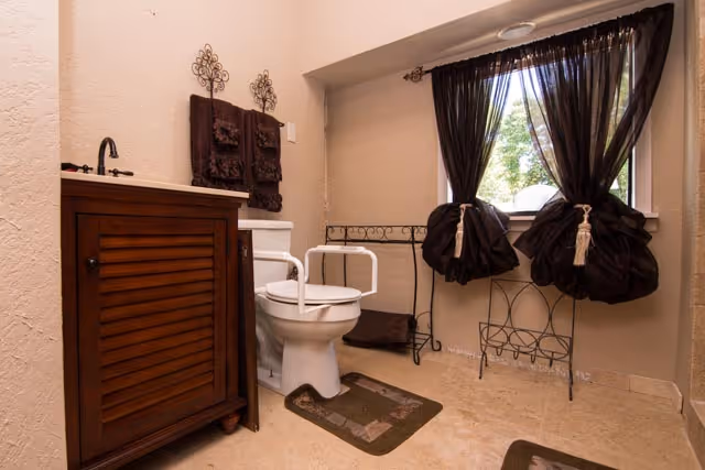 A bathroom with a white toilet equipped with safety rails, a wooden vanity with a sink, brown towels hanging on the wall, decorative wrought iron accents, and a window with dark curtains tied in the middle allowing natural light to enter.