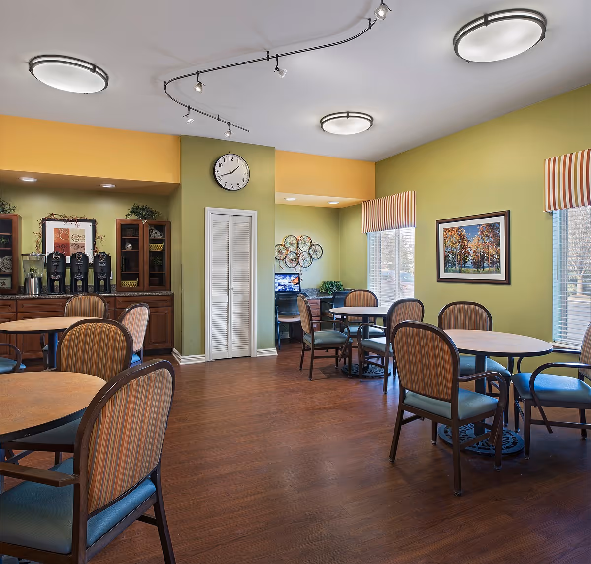 A bright and clean dining area in a senior living facility with round tables and striped cushioned chairs. The walls are painted green and yellow, decorated with framed artwork and a clock. There is a counter with coffee dispensers and cabinets, and a small computer workstation near the windows with striped valances.