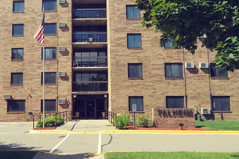 Front exterior view of a multi-story brick building with several windows and balconies. An American flag is flying on a flagpole near the entrance. There is a sign in front of the building that reads 'TALHRIM'. The area is landscaped with bushes and trees, and there is a paved driveway and sidewalk leading to the entrance.