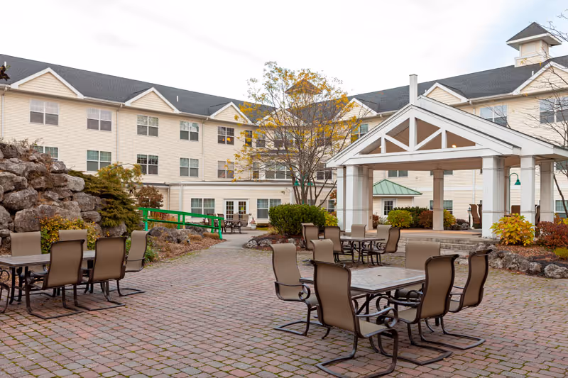 Outdoor patio area at Legacy at Cranberry Landing featuring multiple tables with chairs on a brick-paved surface, surrounded by landscaping with rocks, bushes, and trees, and a large building with multiple windows in the background.