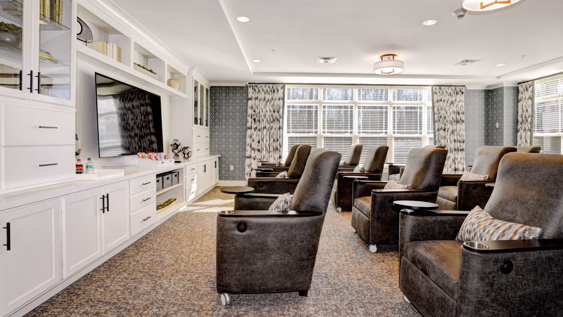 A bright and spacious lounge area with multiple comfortable brown leather recliner chairs arranged in rows facing a large flat-screen TV mounted on a white built-in cabinet wall. The room has large windows with patterned curtains allowing natural light to fill the space, and the carpet has a textured design. Decorative items and books are displayed on the shelves above the TV.