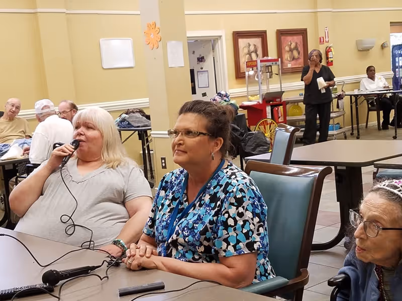 Several residents and staff seated at tables in a communal dining/activity room, one woman speaking into a microphone.