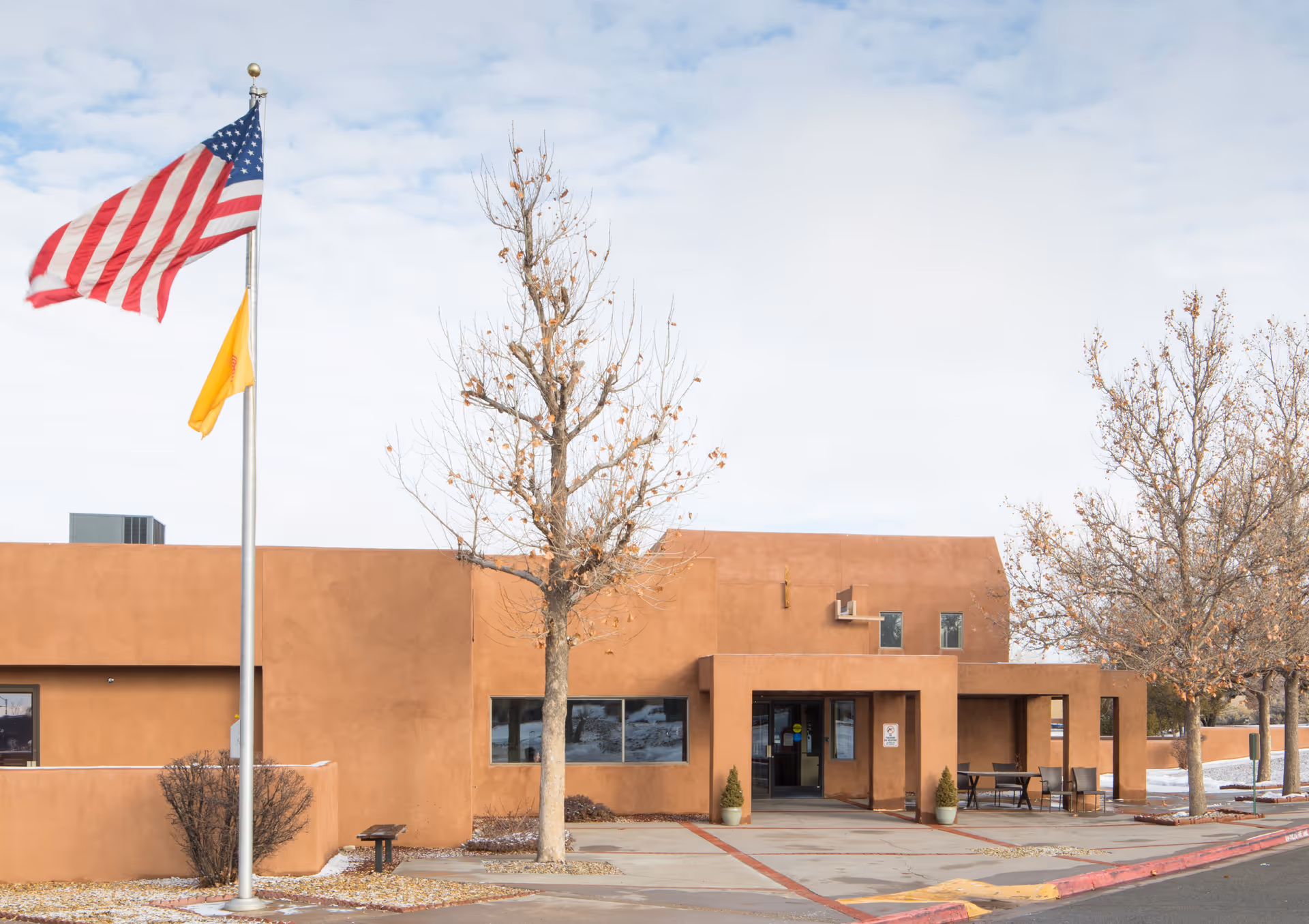 Exterior view of a single-story building with a southwestern architectural style, featuring a flat roof and stucco walls in a warm brown color. In front of the building, there is a flagpole with the American flag and another yellow flag. Leafless trees and some outdoor seating are visible near the entrance. The sky is partly cloudy.