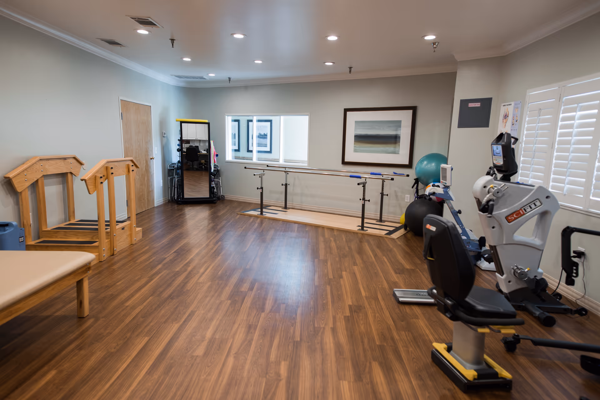 A spacious interior therapy/exercise room with parallel bars, exercise equipment, and wood-look flooring.