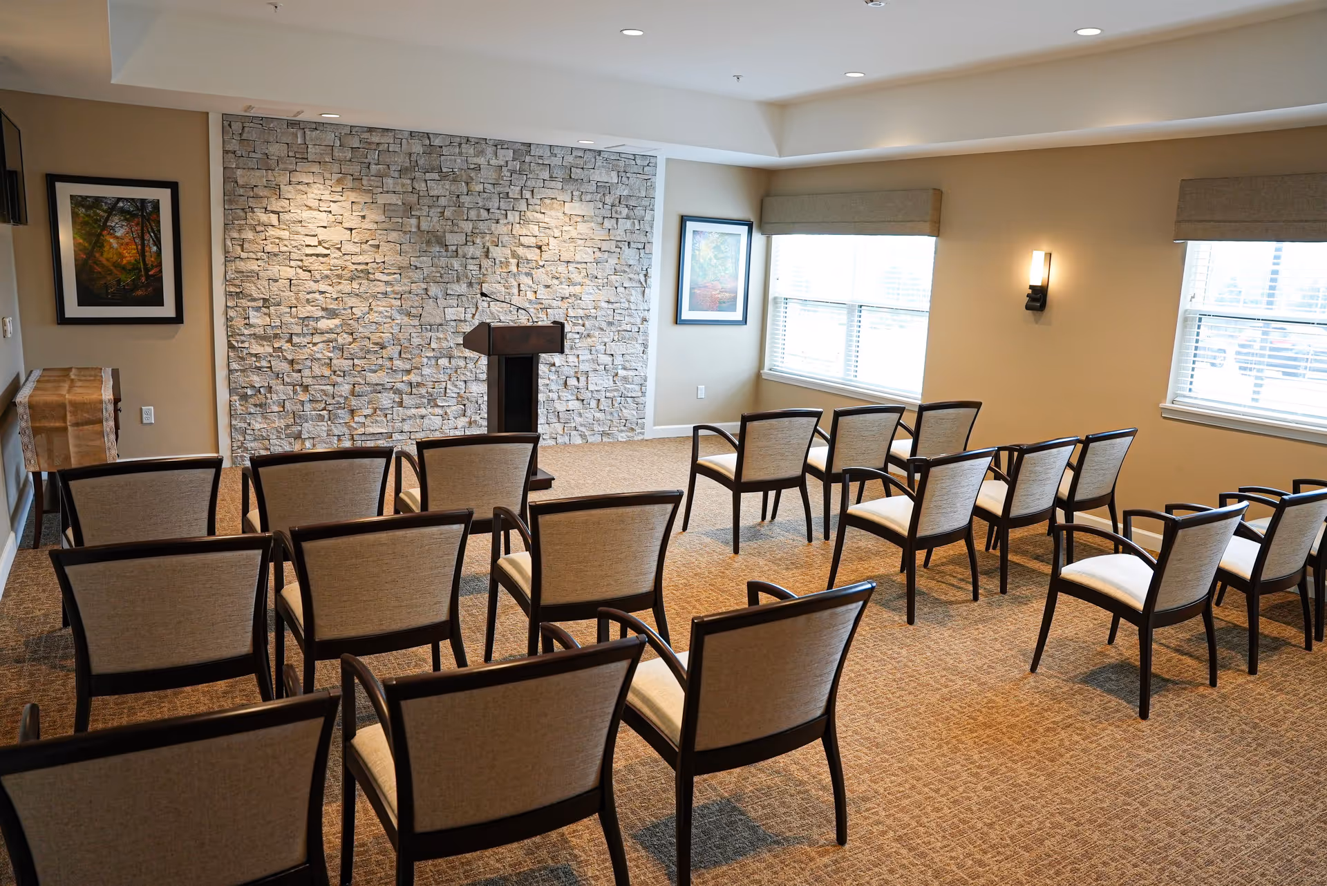 A small conference or meeting room with rows of beige cushioned chairs facing a wooden podium in front of a stone accent wall. The room has beige walls, carpeted floor, two framed pictures on the walls, and two windows with blinds letting in natural light.