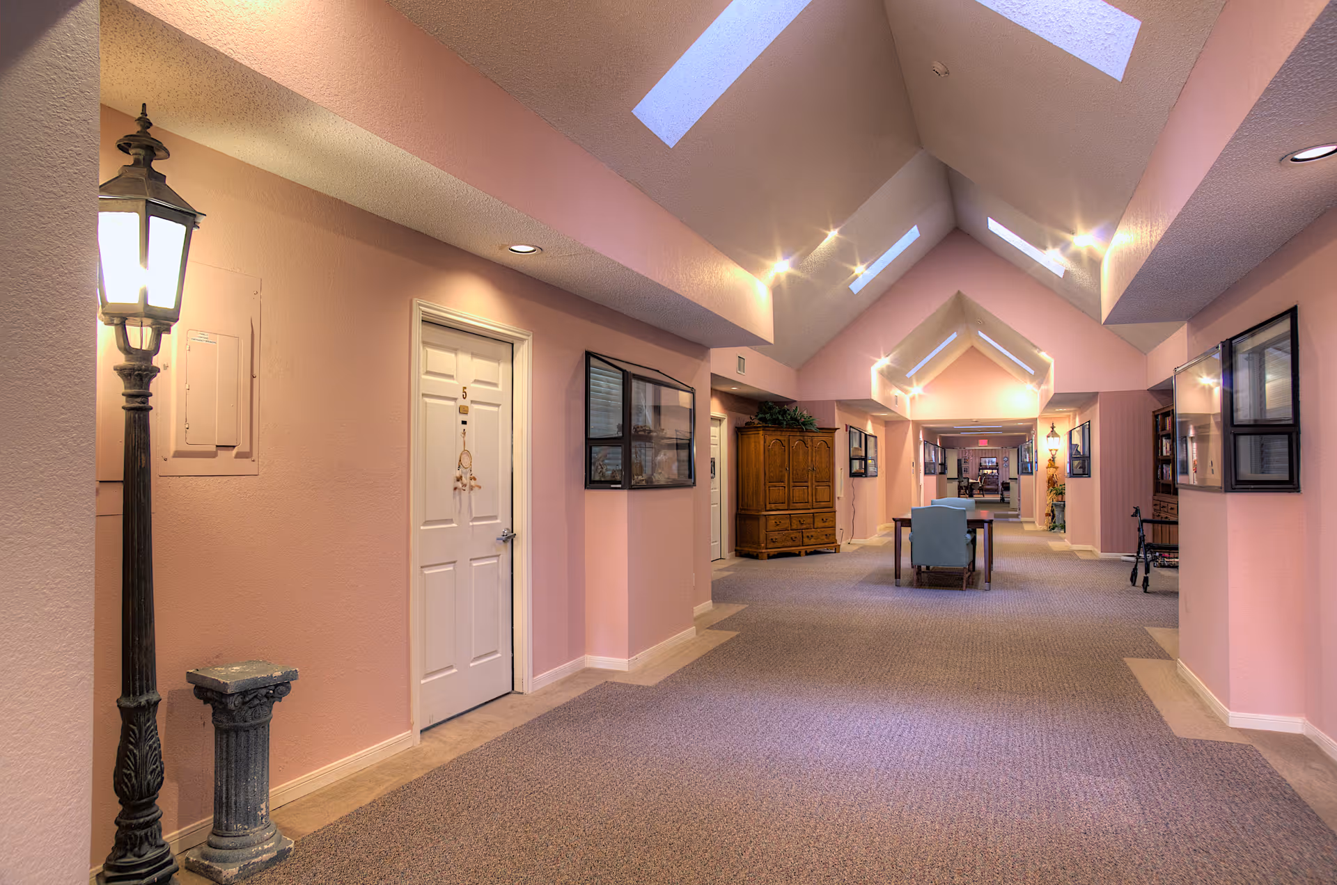 A spacious, well-lit hallway inside Marble Falls Assisted Living with pink walls, a high vaulted ceiling with skylights, and carpeted floors. The hallway features white doors, framed pictures on the walls, a wooden cabinet, a table with chairs, and decorative elements including a vintage-style street lamp and a small pedestal.