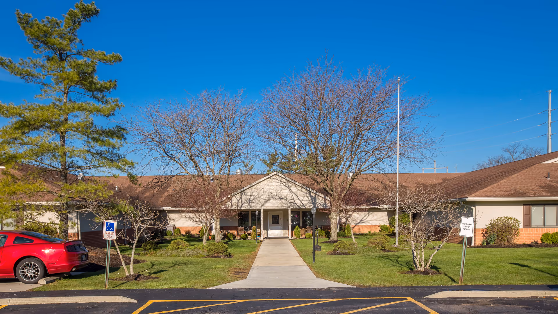 Front exterior view of a single-story care center building with a brown roof, beige walls, and a central entrance. There is a concrete walkway leading to the entrance, surrounded by green grass, small trees without leaves, and some shrubs. A red car is parked in a handicapped parking space on the left side. The sky is clear and blue.