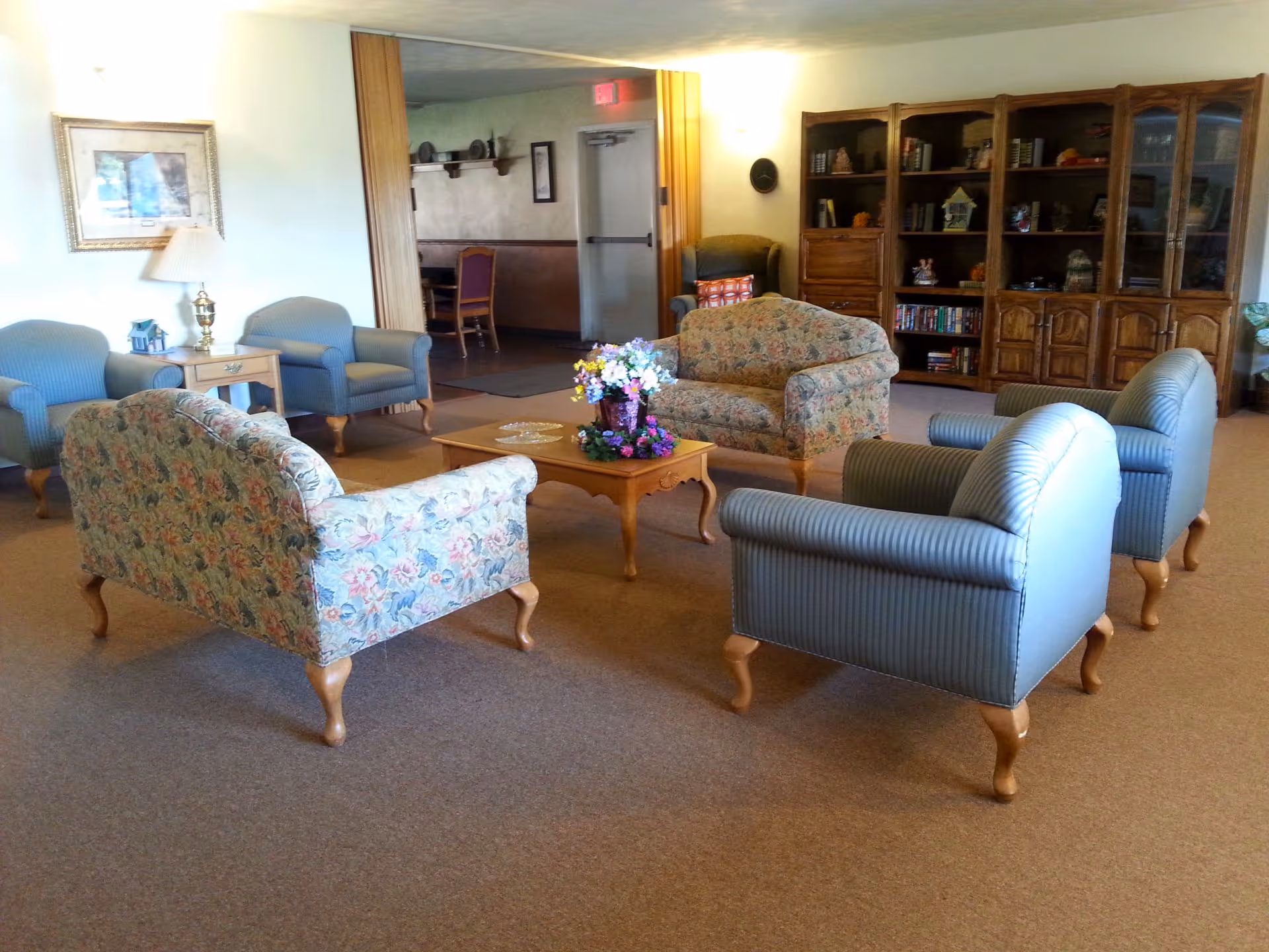 A comfortable sitting room with floral and striped armchairs arranged around a coffee table topped with a flower arrangement and wooden bookshelves along the back wall.