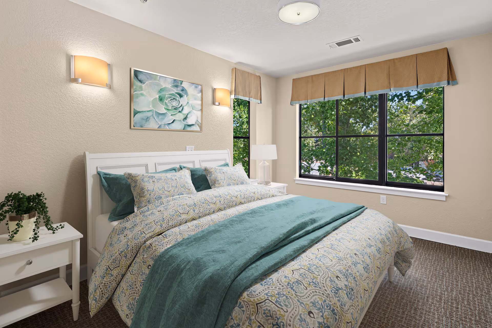 A bright bedroom with a large window showing green trees outside. The room features a white bed frame with patterned bedding in shades of blue and green, two wall-mounted lights above the bed, a framed succulent artwork, a white nightstand with a potted plant, and a table lamp on the other side.