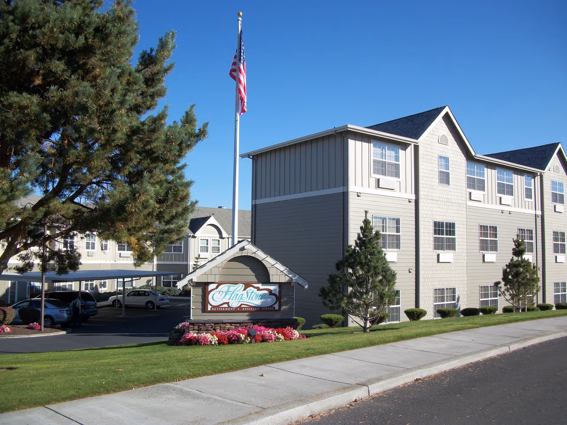 Exterior view of a multi-story senior living facility building with beige siding and multiple windows. There is a well-maintained lawn with small trees and flower beds. A flagpole with an American flag stands next to a sign that reads 'Flagstone Retirement & Assisted Living'. Several cars are parked under a covered parking area to the left of the building.