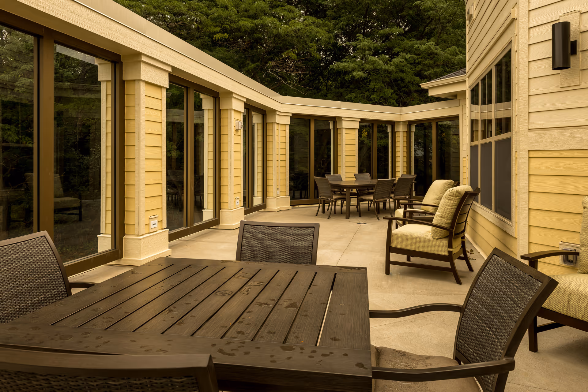 Outdoor patio area with multiple tables and chairs, including cushioned armchairs, adjacent to a building with large windows and surrounded by trees.