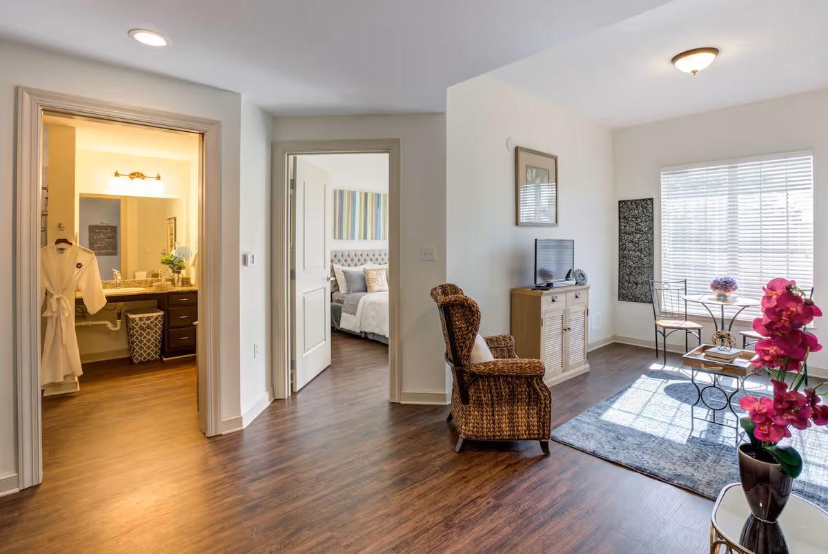 A bright and cozy senior living apartment interior featuring a living area with a wicker armchair, a small TV on a wooden cabinet, a round table with two chairs near a large window with blinds, and a vase with pink flowers on a side table. To the left, there is a bathroom with a lit vanity mirror and a white robe hanging on the door, and a bedroom with a neatly made bed and colorful artwork on the wall.