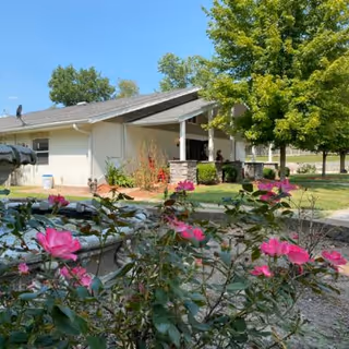 Single-story building with a covered porch surrounded by green trees and a garden with blooming pink flowers in the foreground under a clear blue sky.