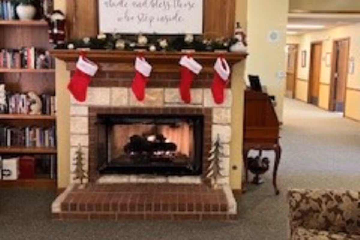 A cozy common room with a stone fireplace decorated with four red Christmas stockings, bookshelves to the left and a hallway to the right.