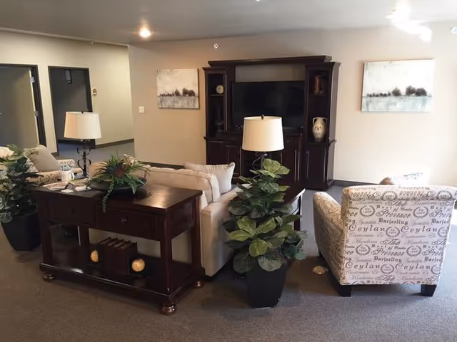 Comfortable seating area with sofas, armchairs, table lamps, potted plants, and an entertainment cabinet in a common room.
