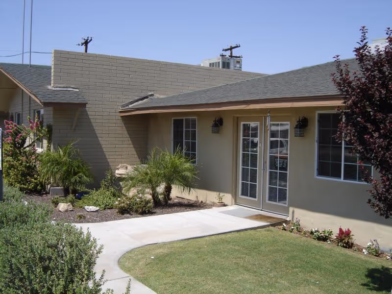 Exterior view of a single-story building with beige walls and a gray shingled roof. There are two windows and a glass door with white frames. The building is surrounded by a well-maintained garden with green shrubs, small palm trees, and flowering plants. A concrete pathway curves through the lawn leading to the door. The sky is clear and blue.