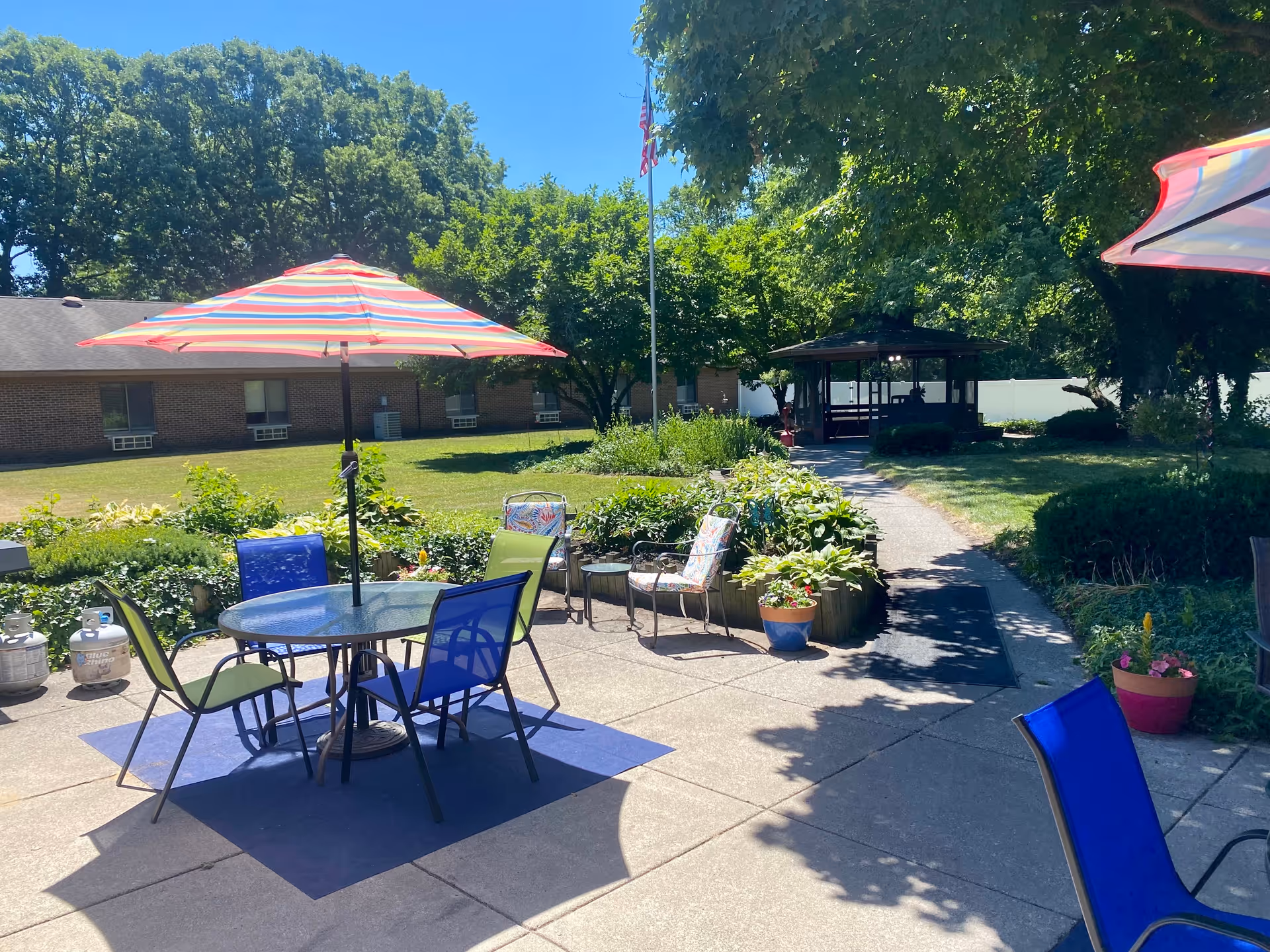 Outdoor patio area with a round glass table and colorful chairs under a striped umbrella. There are plants and flowers in pots and garden beds surrounding the patio. A pathway leads to a gazebo in the background, with trees and a brick building visible under a clear blue sky.