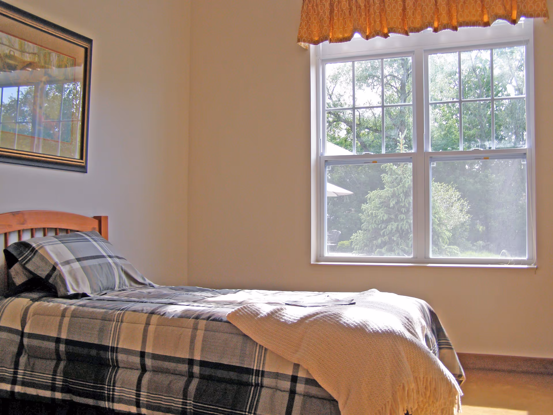 A bright bedroom with a single bed covered in a plaid blanket and a pillow. A cream-colored throw blanket is draped over the foot of the bed. There is a large window with a valance, letting in natural light and showing green trees outside. A framed picture hangs on the wall above the bed.