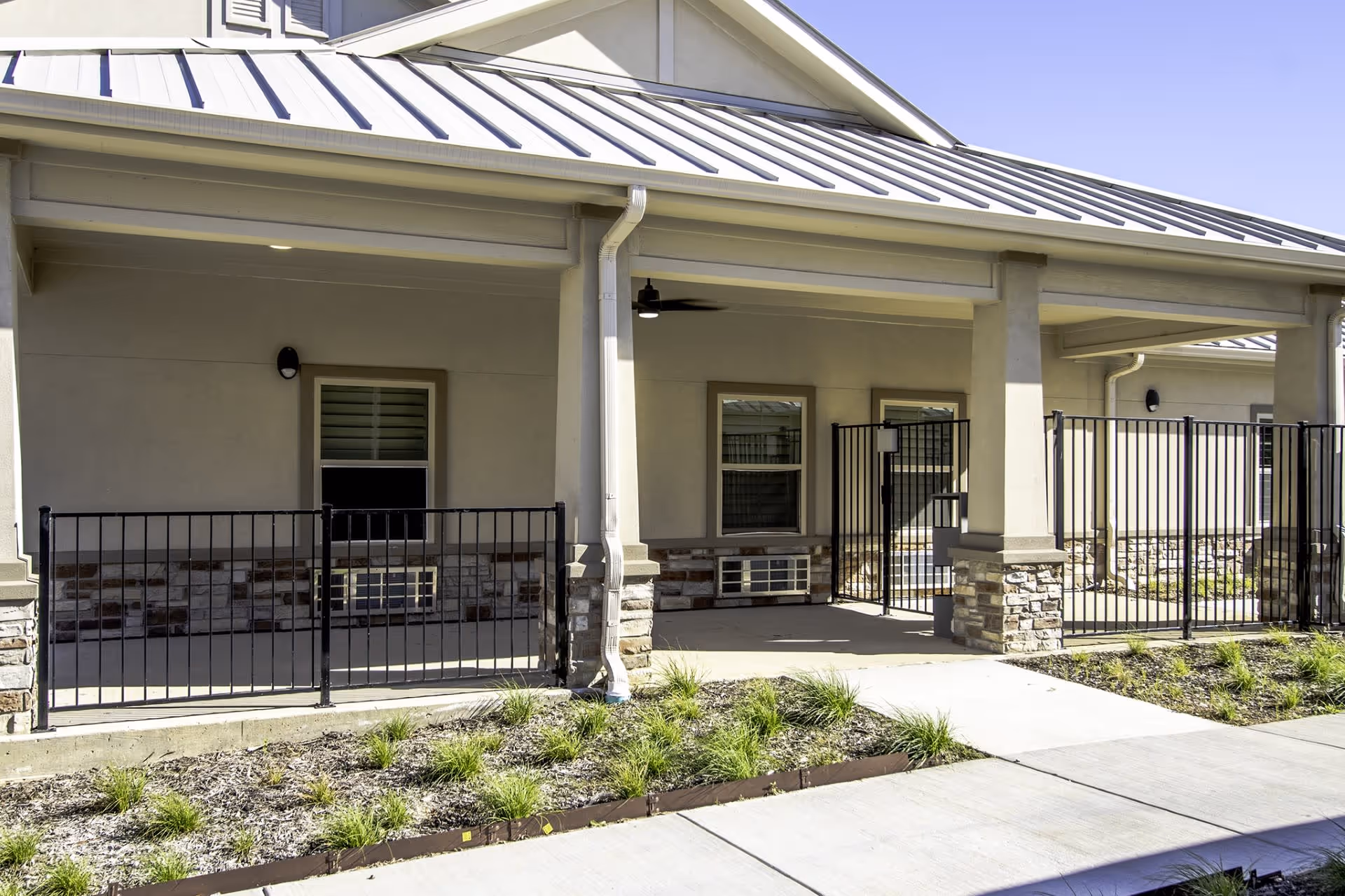 Exterior view of a single-story building with a metal roof and beige walls. The building features a covered porch area with stone pillars and black metal fencing enclosing small patio spaces. There are windows with air conditioning units beneath them and a concrete walkway in front with some landscaping including small green plants.