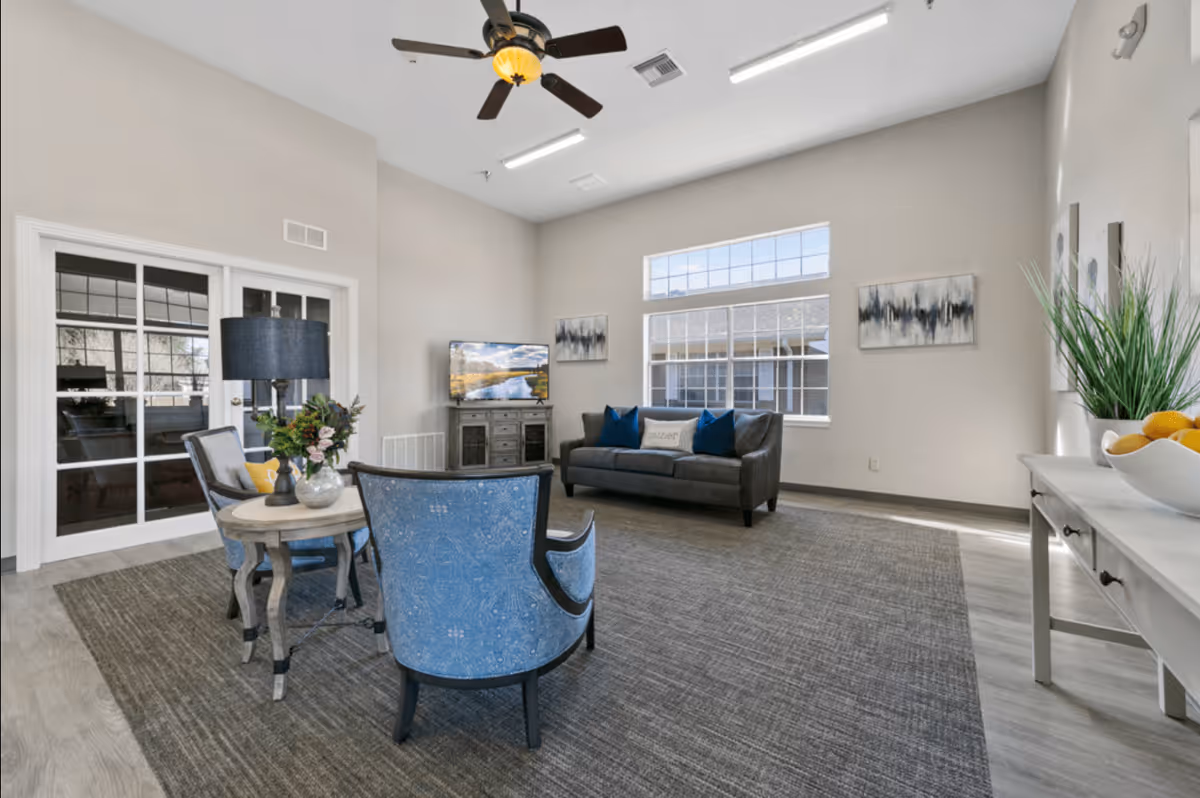 A bright and spacious living room with a gray sofa adorned with blue and white pillows, two blue upholstered chairs around a small round table with a lamp and flower arrangement, a flat-screen TV on a wooden stand, large windows letting in natural light, a ceiling fan, and a white console table with a bowl of oranges and a green plant.