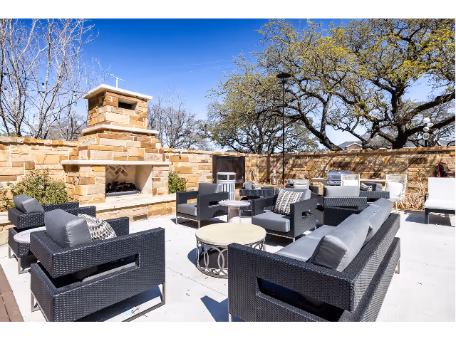 Sunny outdoor courtyard with black wicker lounge seating arranged around round tables and a stone fireplace beneath trees.