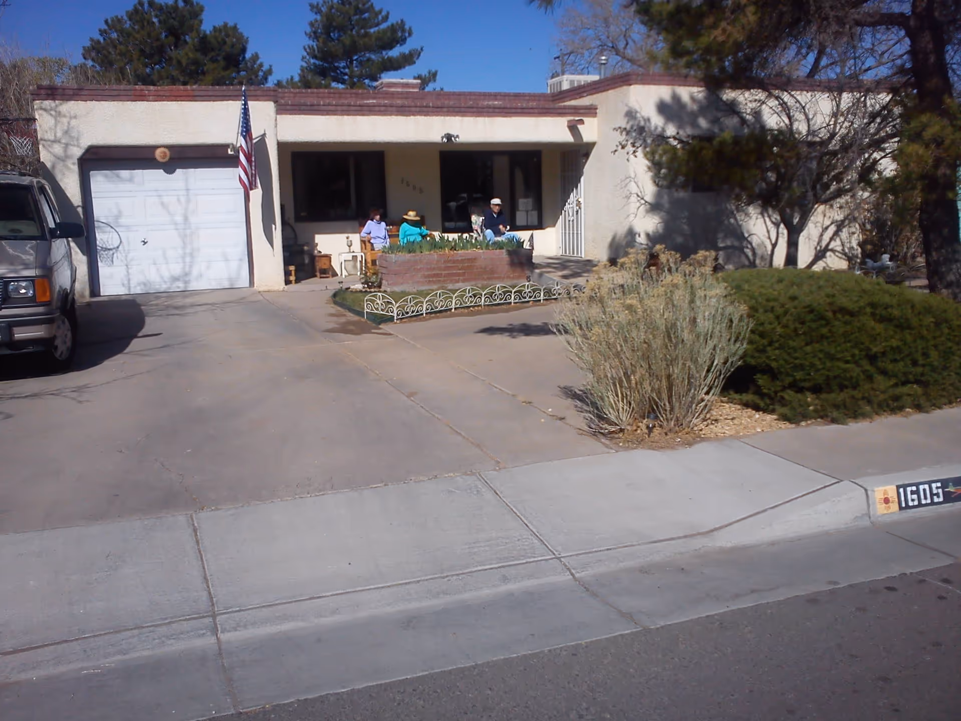 Front exterior view of a single-story residential building with a garage, driveway, and a small garden area. Three people are sitting on the porch near the entrance. There is an American flag mounted near the garage door. The house number 1605 is visible on the curb.