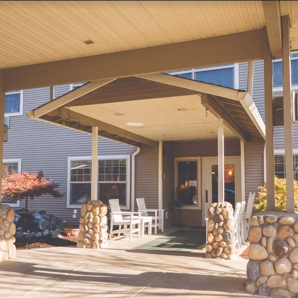 Covered main entrance with a pitched canopy supported by stone pillars, white chairs on the porch, and double glass doors.
