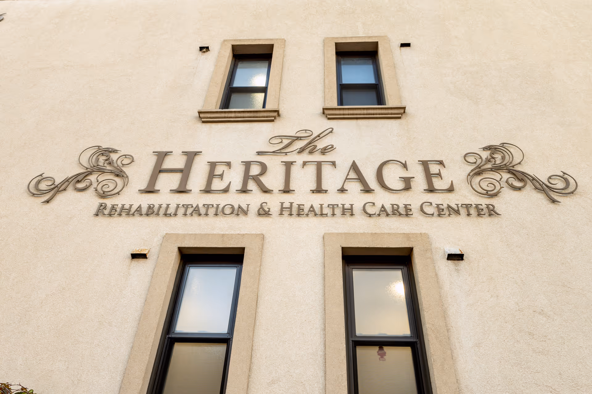 Exterior wall of a building with the sign 'The Heritage Rehabilitation & Health Care Center' mounted between two sets of windows.