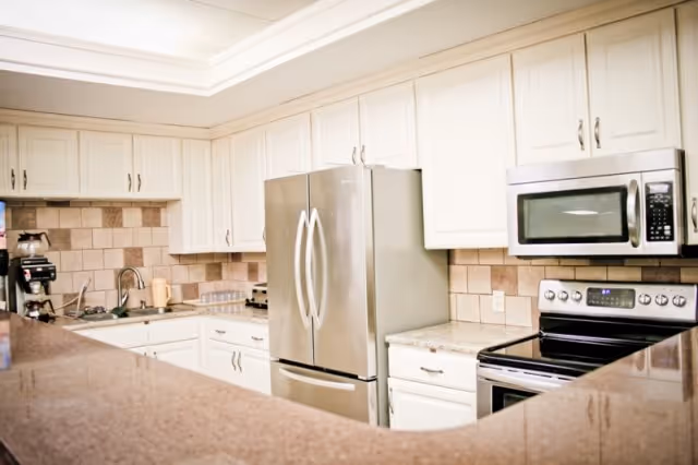 A modern kitchen with white cabinets, a stainless steel refrigerator, microwave, and electric stove. The countertops are a polished brown stone, and there is a tiled backsplash in beige tones. A coffee maker and some kitchen utensils are visible on the counter.