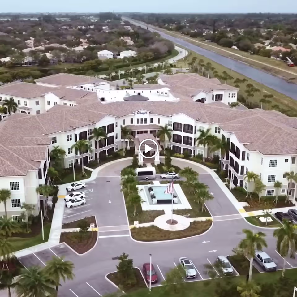 Aerial view of Allegro Senior Living facility in Parkland, FL, showing a large, multi-wing building with a circular driveway and a central fountain with an American flag. The building is surrounded by palm trees, parking spaces, and a canal running parallel to a road in the background.