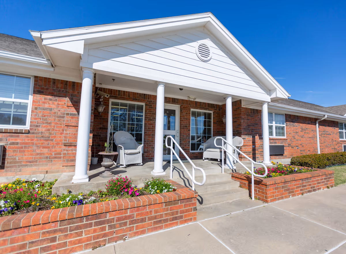 Front entrance of a brick building with a covered porch supported by white columns, steps with handrails, patio chairs, and flower beds.