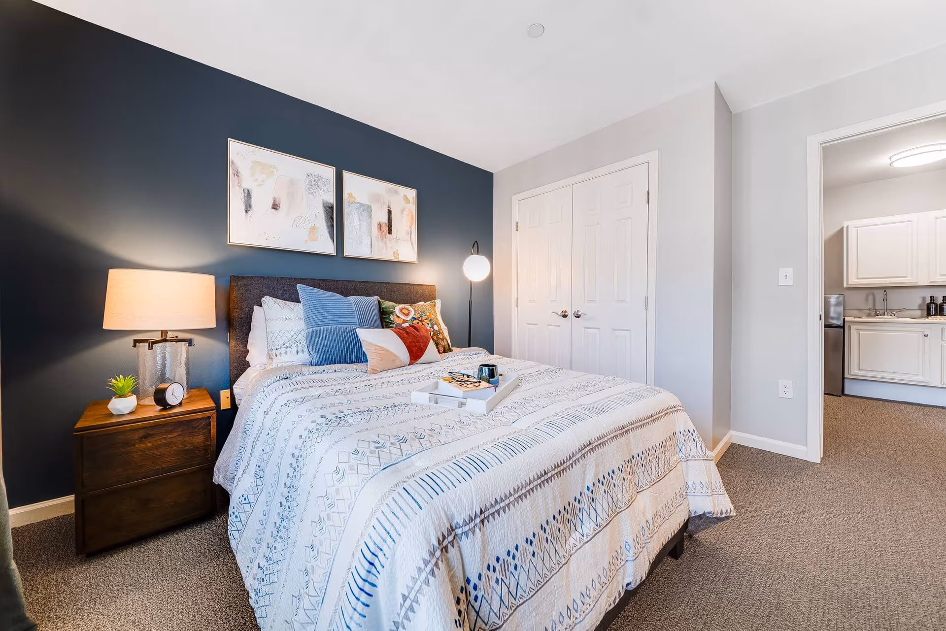 A cozy bedroom with a neatly made bed featuring patterned bedding and multiple decorative pillows. There is a wooden nightstand with a lamp, a small plant, and a clock. The wall behind the bed is painted dark blue and adorned with two abstract art pieces. A floor lamp with a round white shade stands near a white double-door closet. An open doorway reveals a glimpse of a kitchen area with white cabinets and a sink.