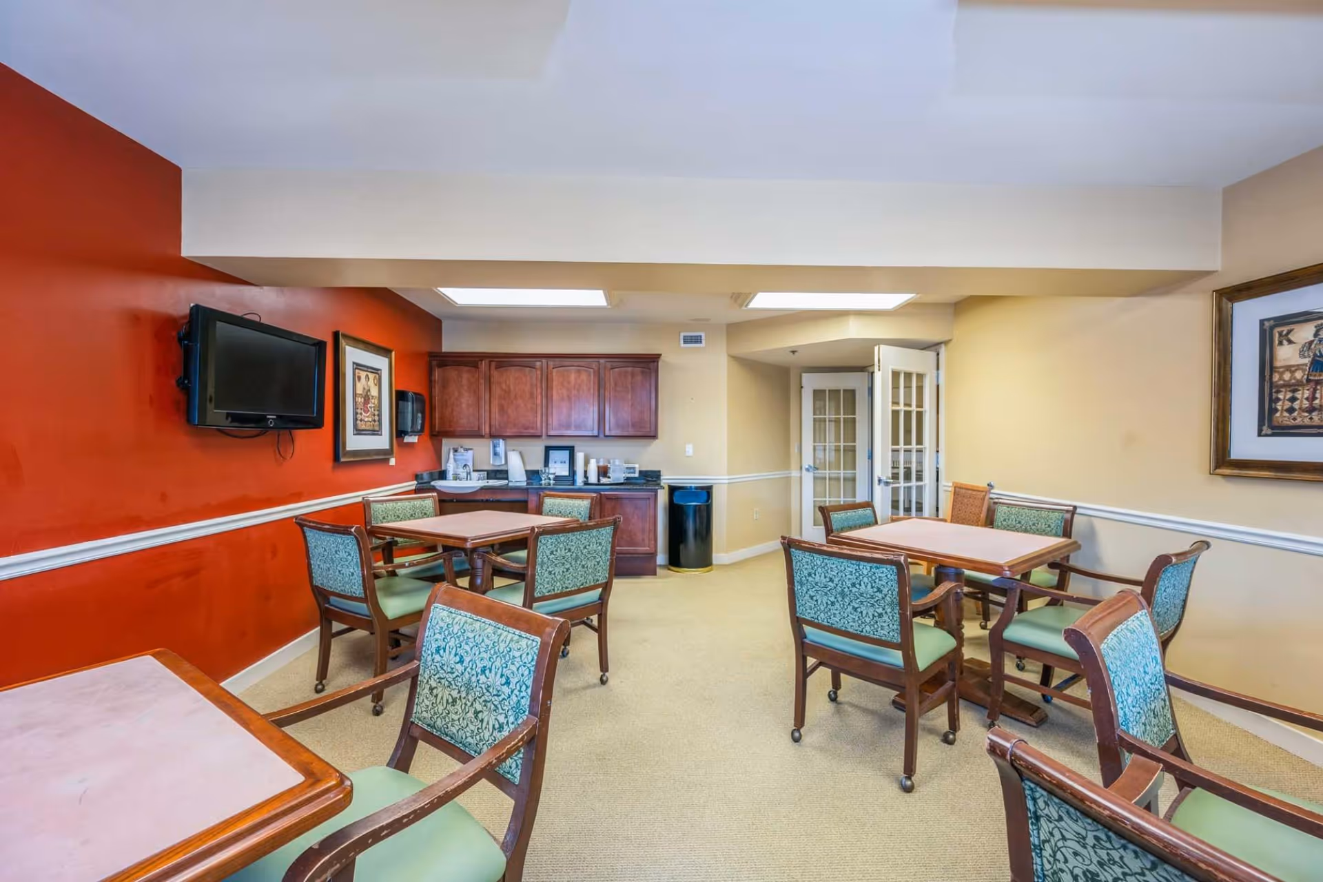 A small dining area with several square wooden tables and green cushioned chairs with patterned backs. The room has a red accent wall with a mounted flat-screen TV and framed artwork. There are wooden cabinets and a countertop with various items against a beige wall. The floor is carpeted, and there are double glass doors in the background.