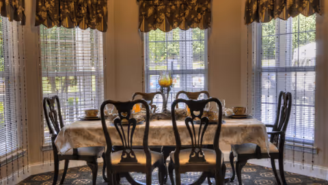 A dining room with a rectangular table covered with a patterned tablecloth, surrounded by six dark wooden chairs with decorative backs. The room has large windows with blinds and floral valances, allowing natural light to brighten the space. A decorative centerpiece with candles is placed on the table.
