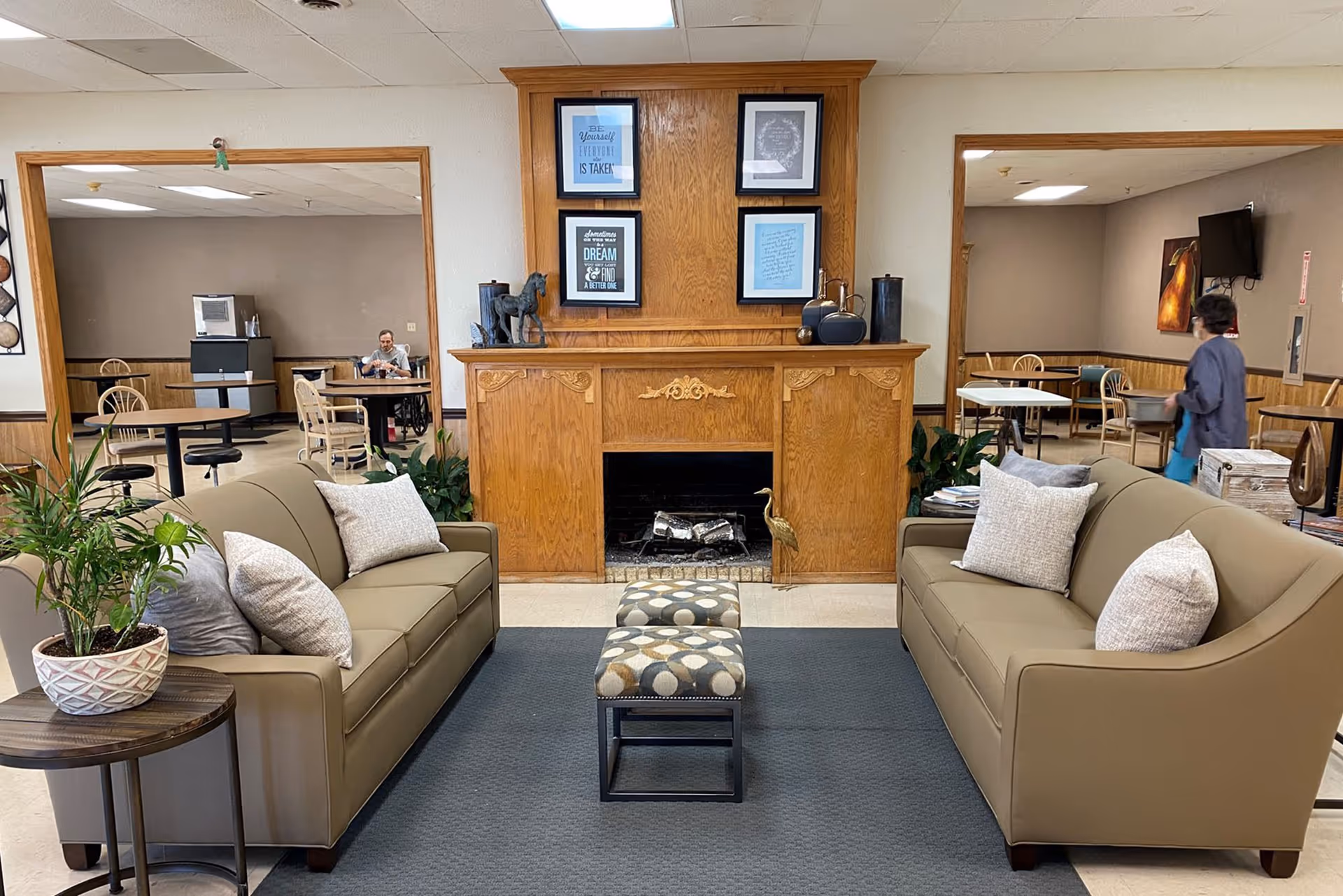 Communal lounge with two sofas facing a wooden fireplace and ottoman, with a dining area and people visible in the background.
