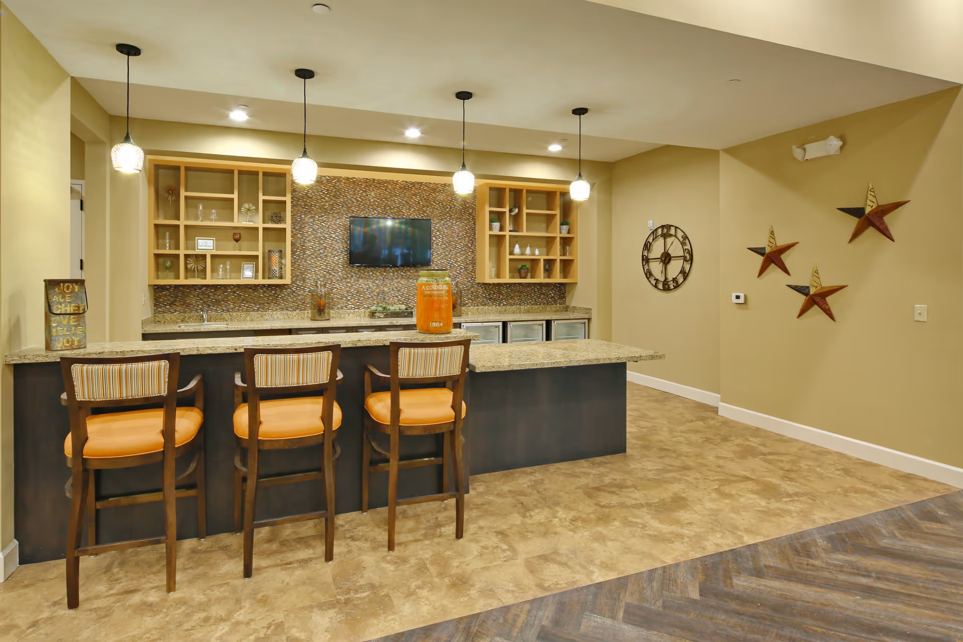 Interior view of a modern kitchen area with a granite countertop island and three wooden bar stools with orange cushions. The back wall features mosaic tile backsplash, open shelving with decorative items, a mounted flat-screen TV, and three hanging pendant lights. The adjacent wall has a large clock and three decorative star wall hangings.