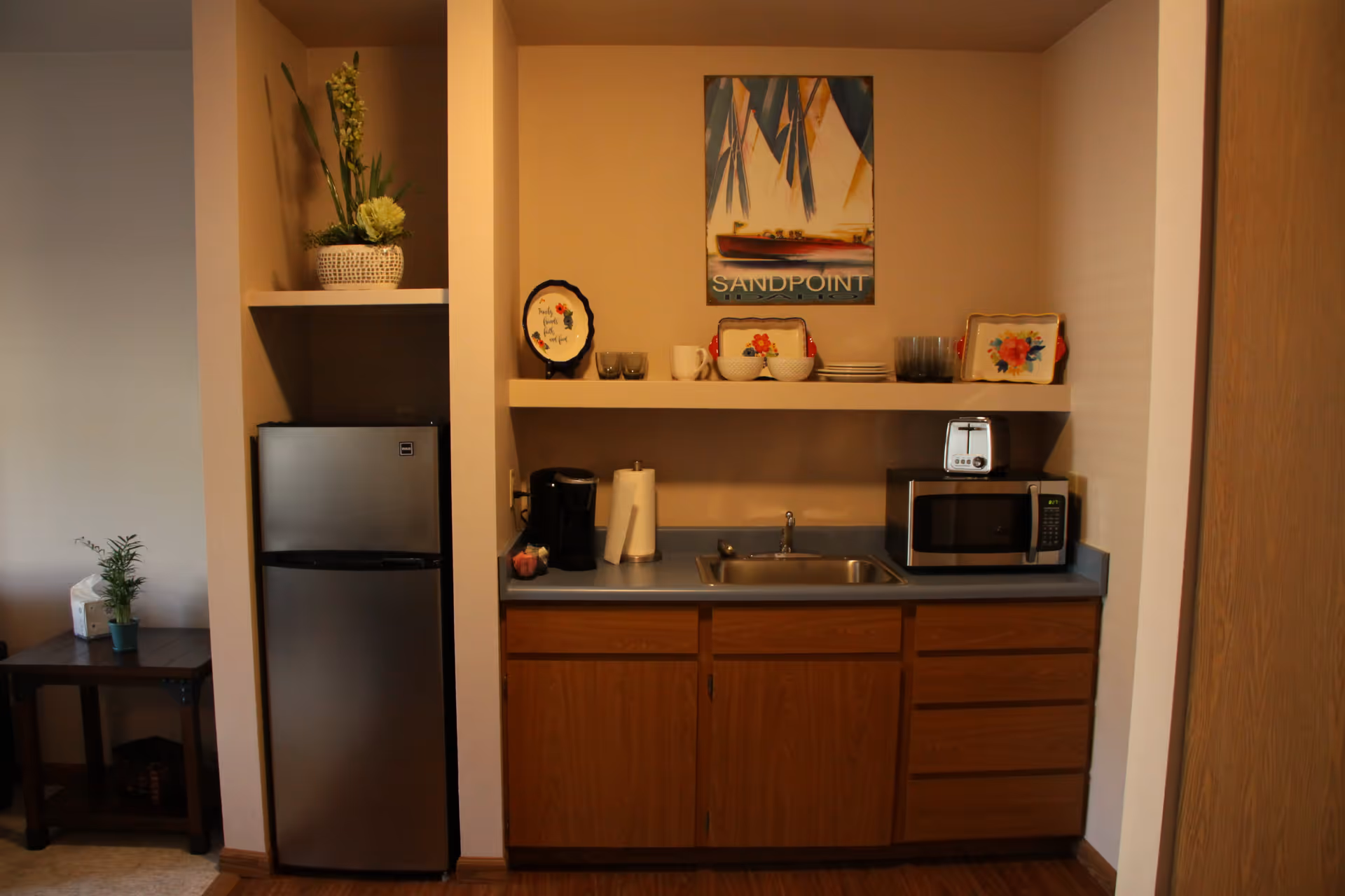 A small kitchen area with a stainless steel refrigerator on the left, wooden cabinets below a countertop with a sink in the middle. On the countertop, there is a coffee maker, paper towel holder, and a microwave with a toaster on top. Above the countertop is a shelf holding plates, bowls, mugs, and decorative trays. A framed picture of a sailboat with the word 'SANDPOINT' hangs on the wall above the shelf. To the left of the refrigerator is a small table with a potted plant and a tissue box.