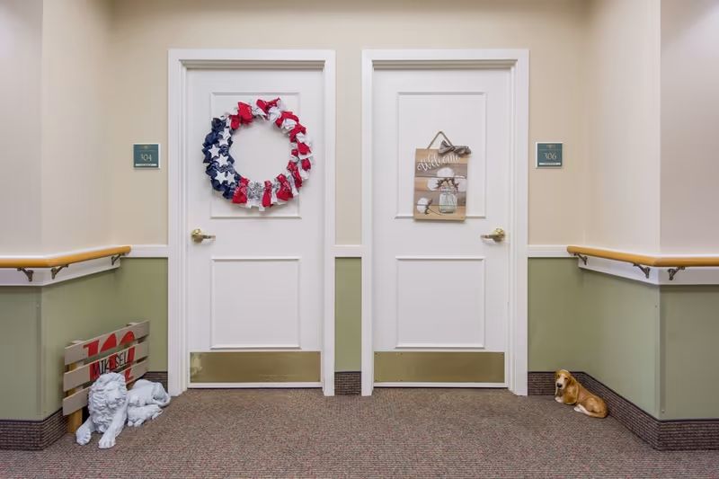 A hallway in a senior living facility with two white doors labeled 304 and 306. Door 304 has a red, white, and blue wreath, and door 306 has a wooden welcome sign with cotton flowers. The hallway has green and beige walls with wooden handrails. There are two decorative dog statues on the floor near the doors, one white and one brown.