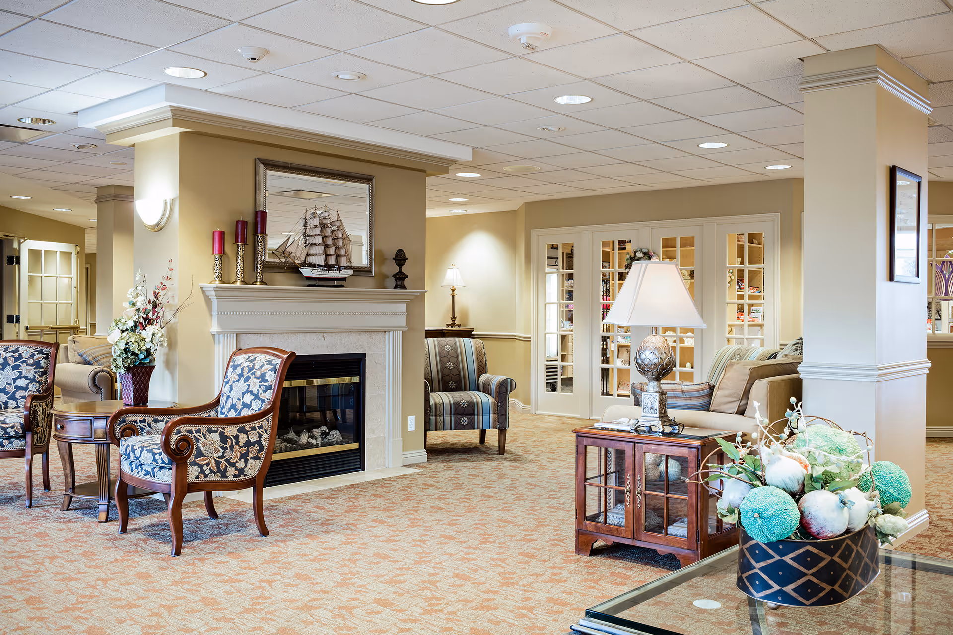 A cozy senior living facility common area with patterned armchairs, a fireplace adorned with a model ship and candles, a side table with a lamp, and a floral arrangement on a glass table. The room features beige walls, carpeted floors, and French doors leading to another room.