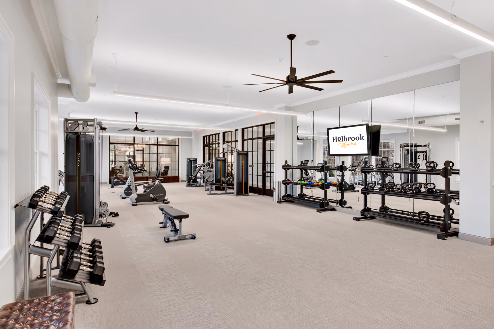Bright, spacious fitness room with weight racks, cardio machines, mirrors and a TV displaying 'Holbrook'.