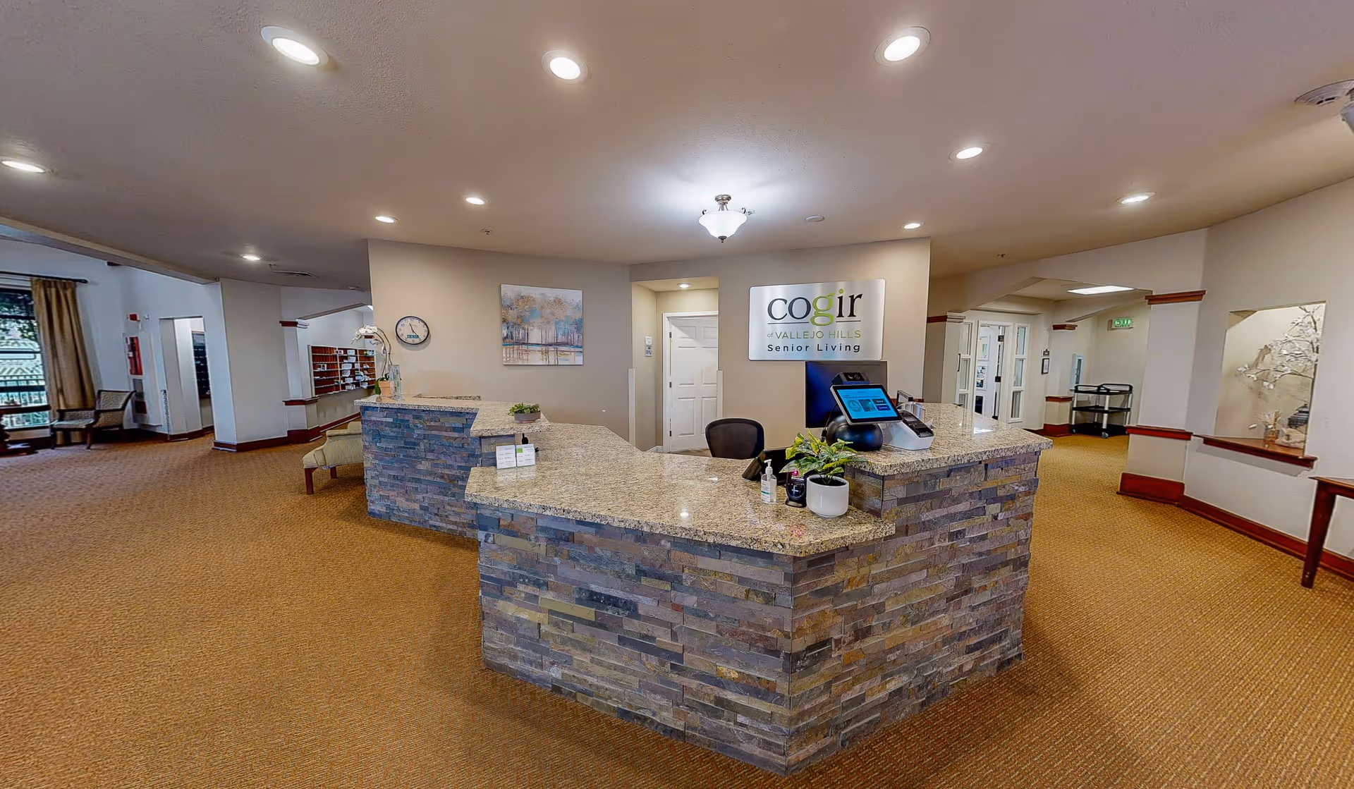 Reception area of Cogir of Vallejo Hills Senior Living with a stone and granite front desk, a computer monitor, potted plants, a wall clock, and a sign displaying the facility's name. The area is well-lit with ceiling lights and has carpeted flooring with beige walls and wooden trim.
