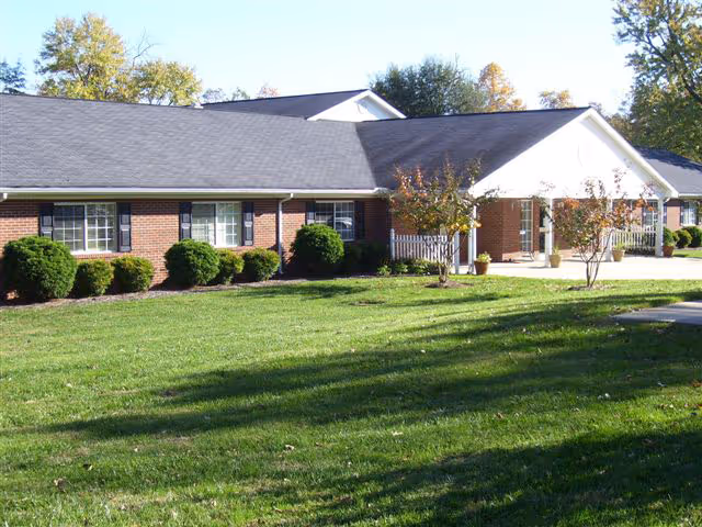 Single-story brick memory care building with a covered entrance, shrubs, and a large grassy lawn.