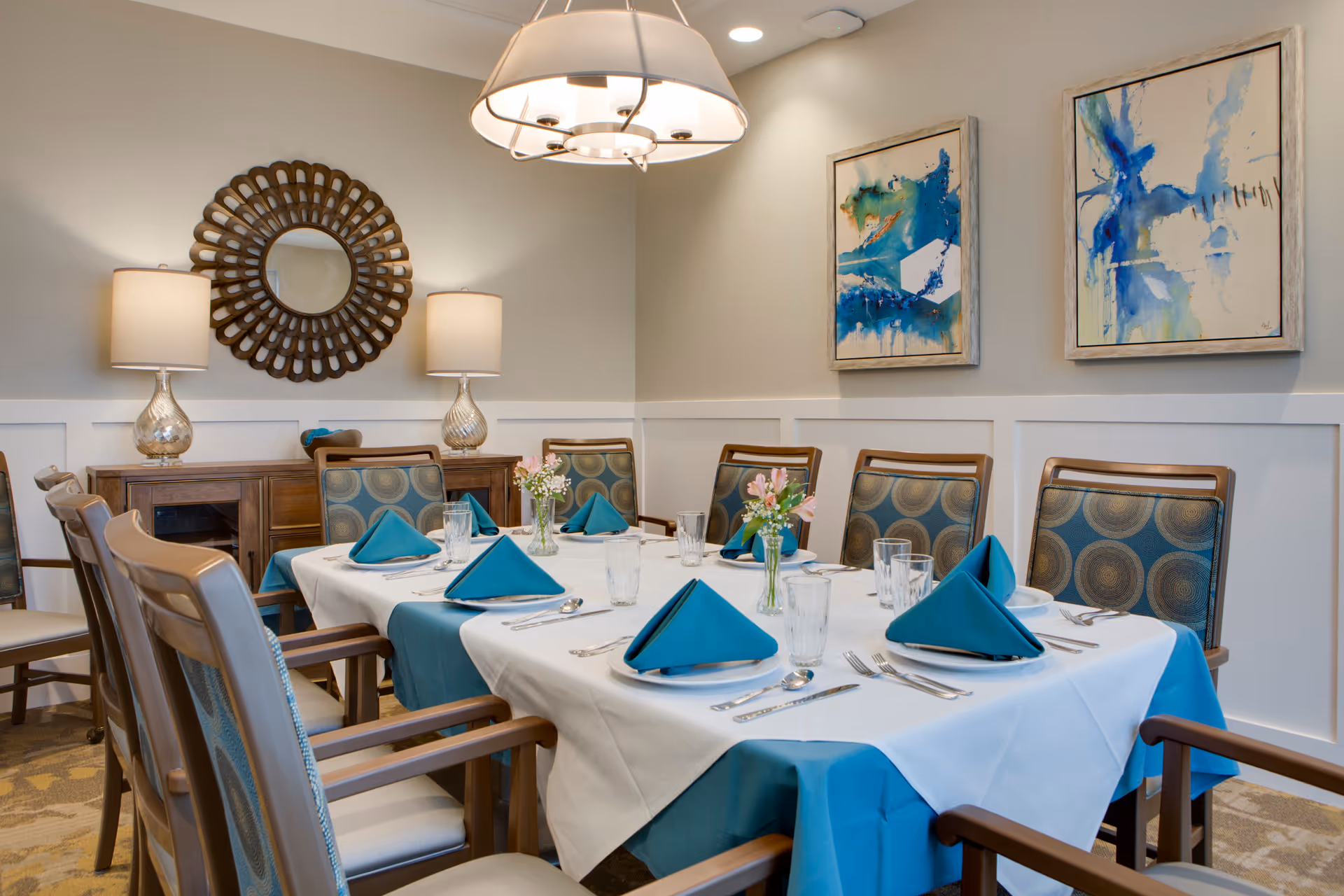 A dining room with a table set for six people, featuring teal napkins folded on white tablecloths with teal underlays. The chairs have patterned upholstery with circular designs. The room is decorated with two abstract blue paintings on the wall, a round decorative mirror, two table lamps on a wooden sideboard, and a modern ceiling light fixture.