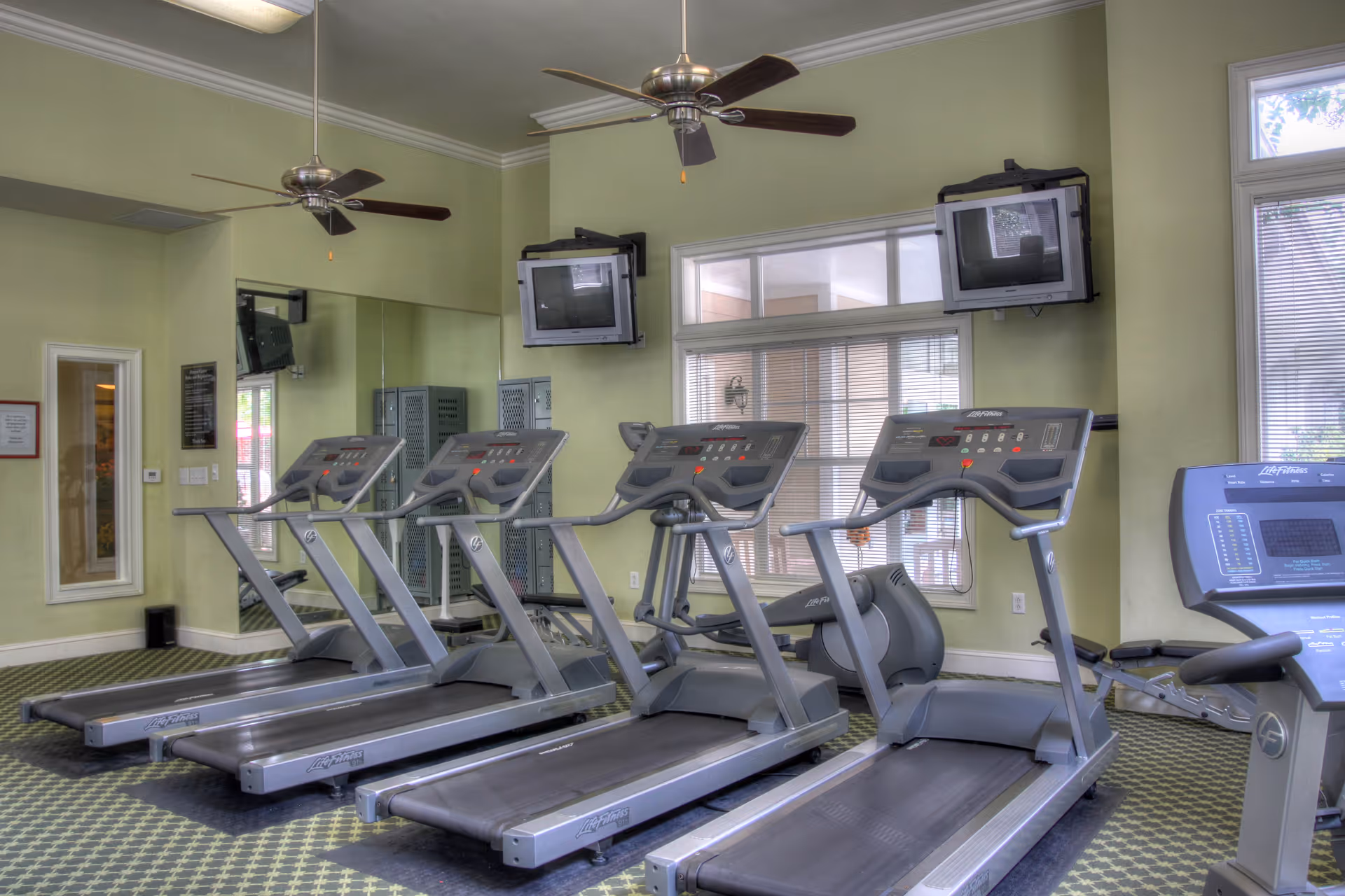 Interior view of a fitness room with four treadmills lined up facing two wall-mounted televisions. The room has light green walls, large windows with blinds, a wall mirror, ceiling fans, and patterned carpet flooring.