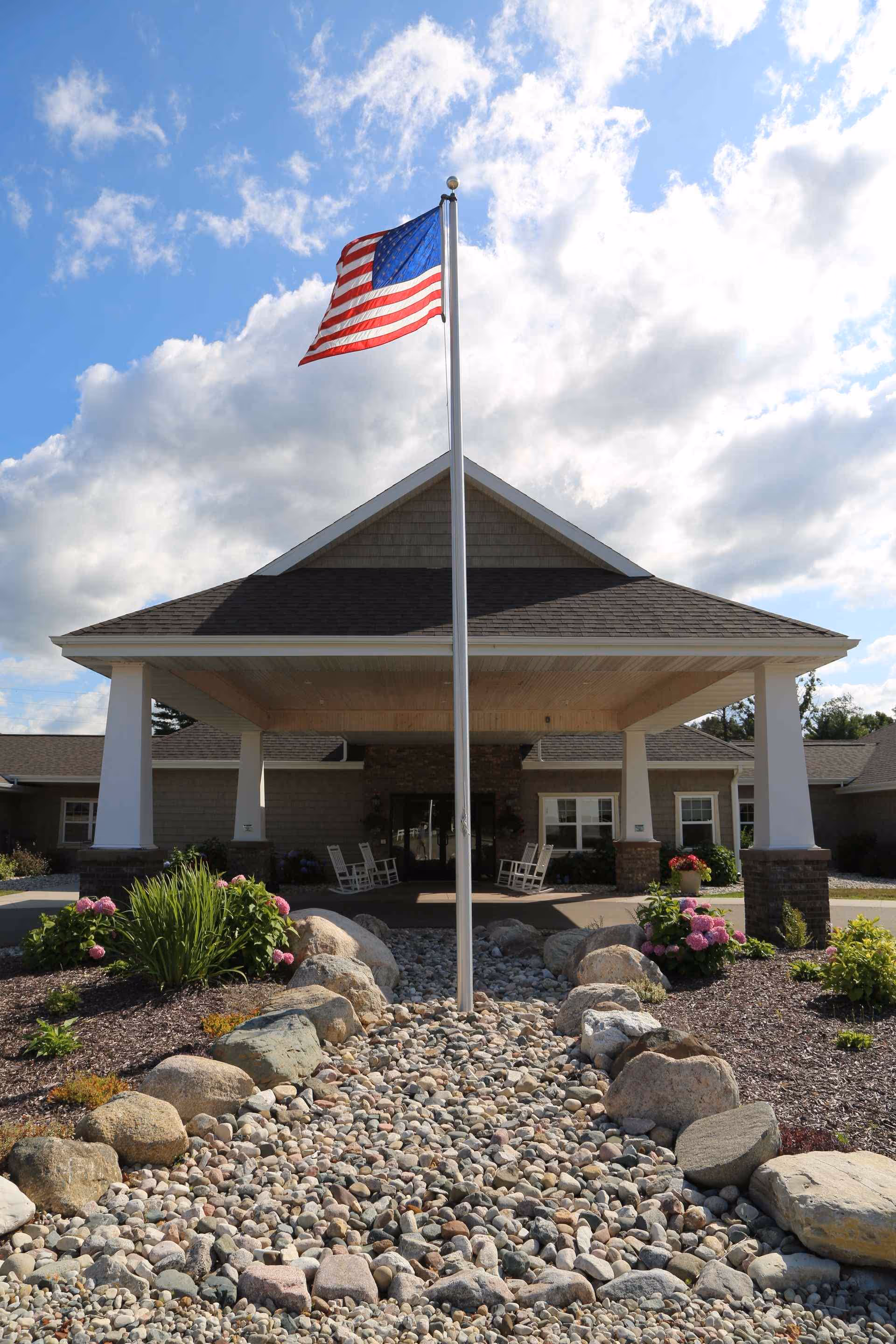 Entrance of a building with a centered flagpole flying an American flag over a covered driveway and landscaped rock beds.