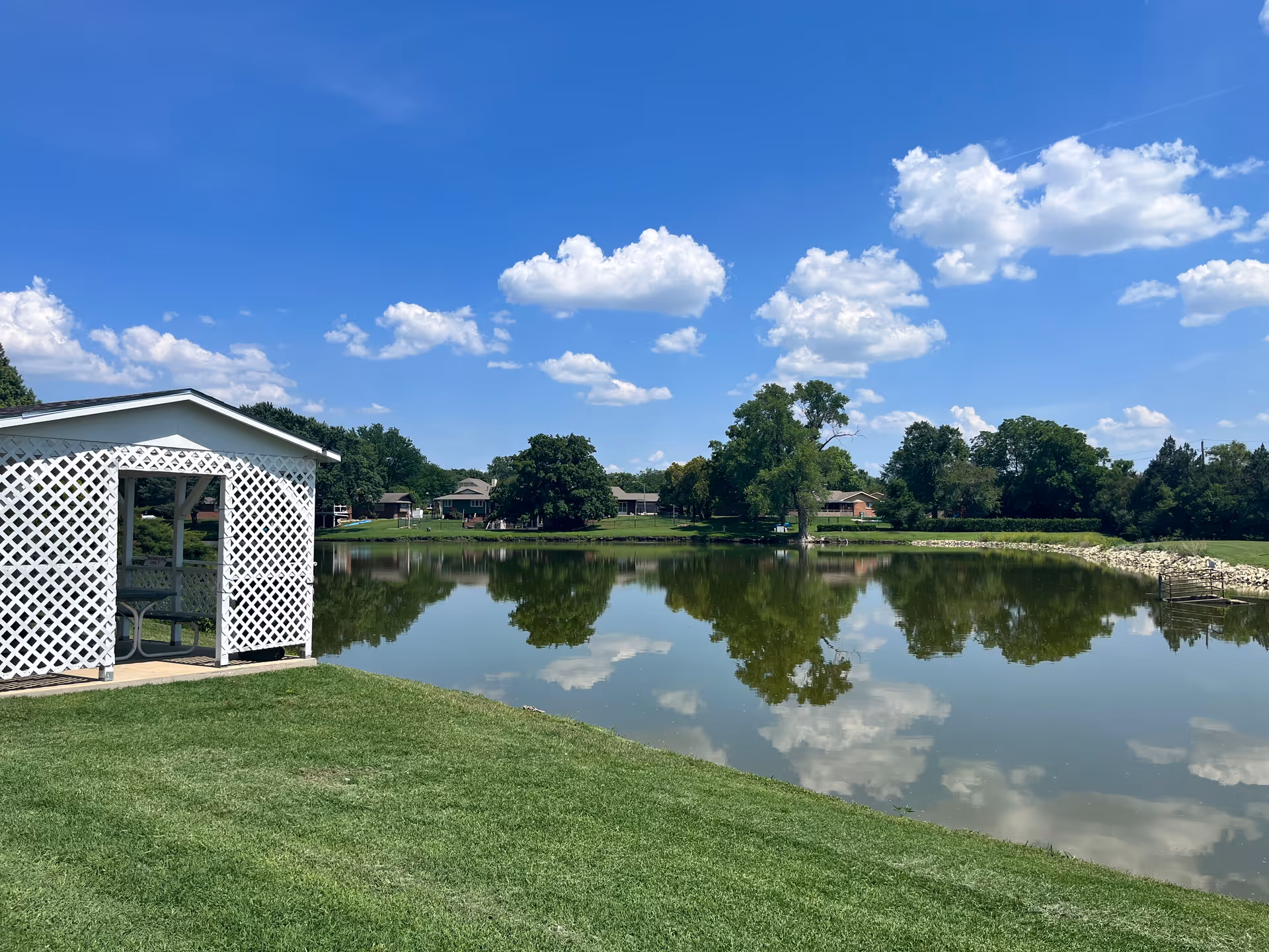Lakeside view showing a white lattice gazebo on a grassy shore with a pond reflecting trees, nearby buildings, and a blue sky with clouds.