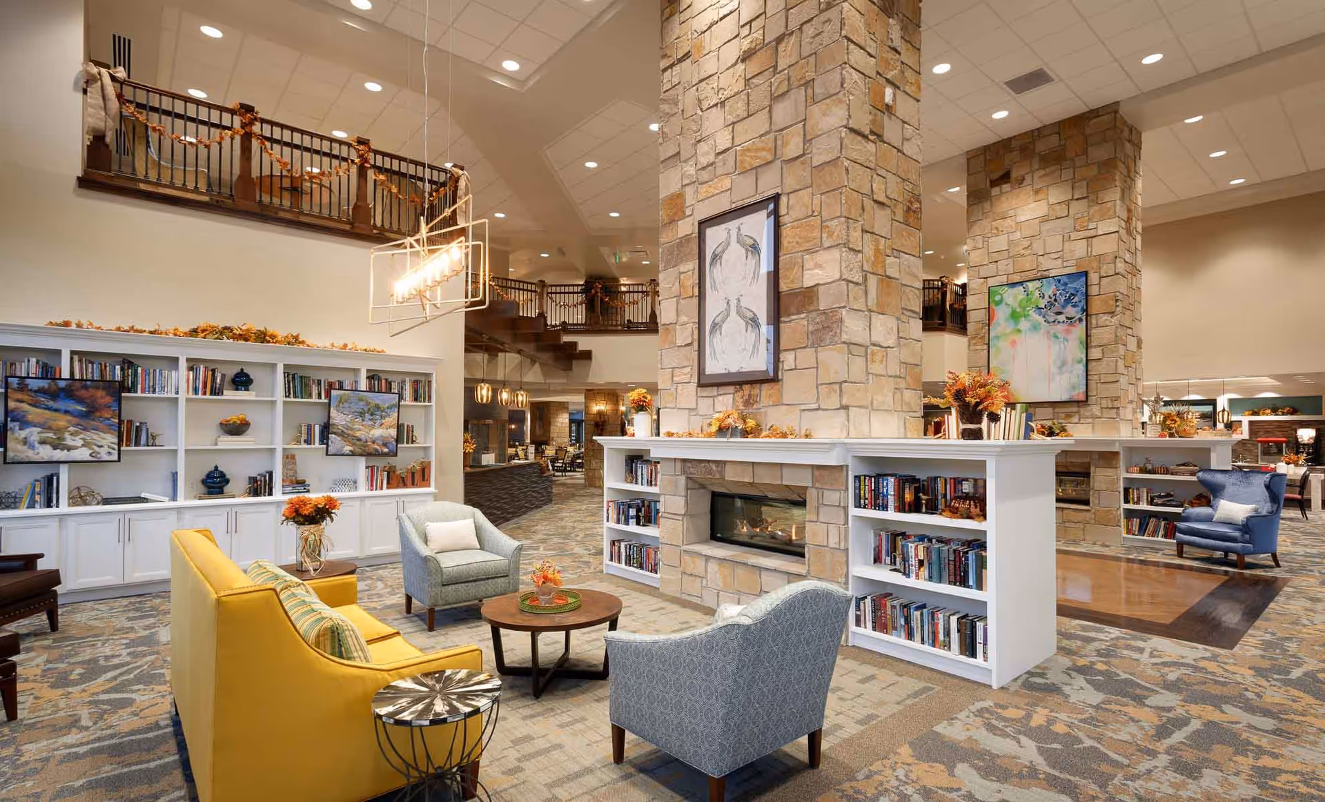 A spacious and well-lit senior living facility common area with a stone fireplace surrounded by built-in bookshelves filled with books. The seating area includes a yellow sofa, two patterned armchairs, and a small round wooden table with a flower arrangement. The room features high ceilings, modern lighting fixtures, and decorative artwork on the walls. There is a second level with a railing decorated with autumn garlands.