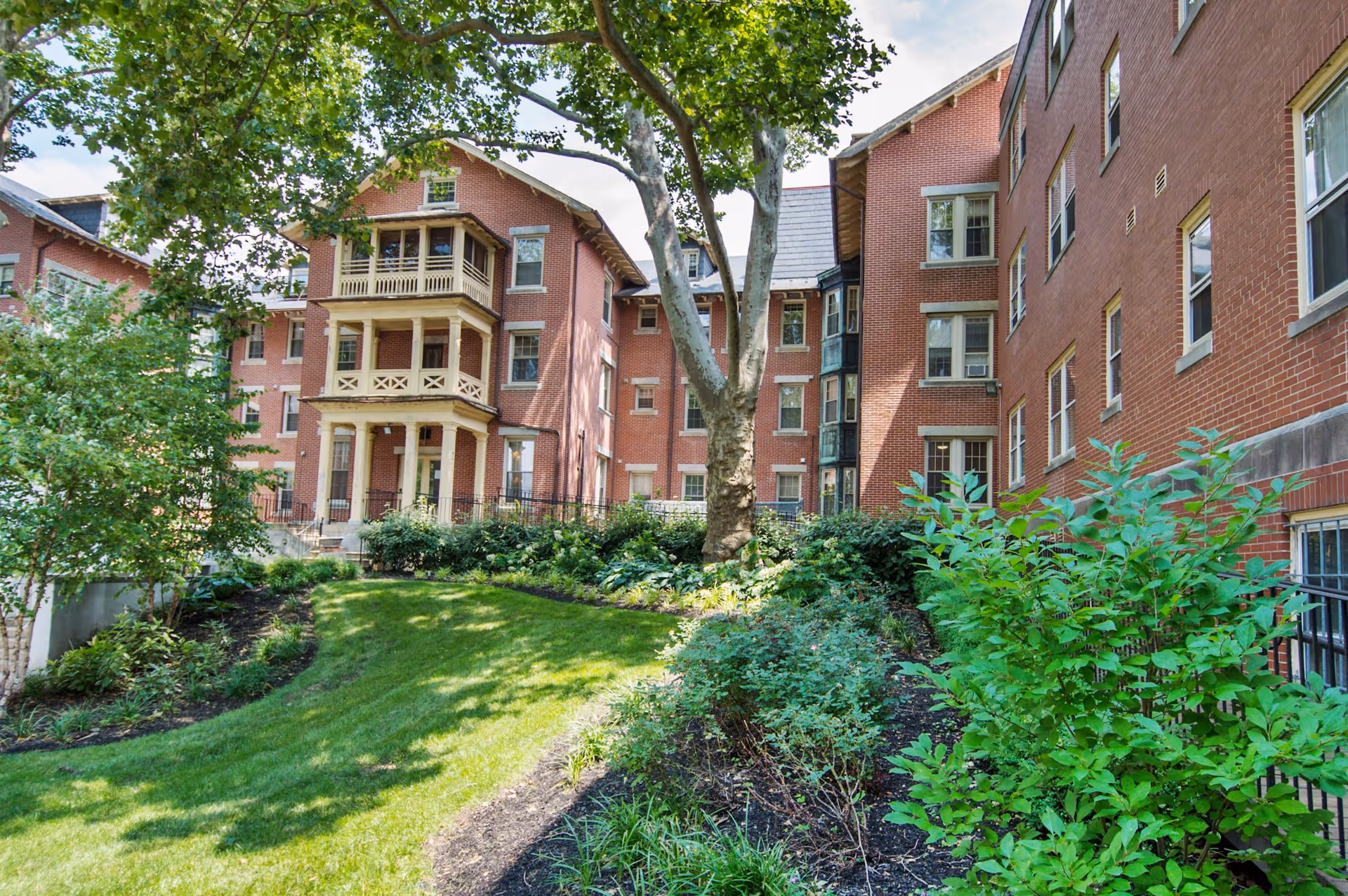 A well-maintained garden area with green grass, shrubs, and trees in front of a large multi-story red brick building with multiple windows and balconies under a partly cloudy sky.
