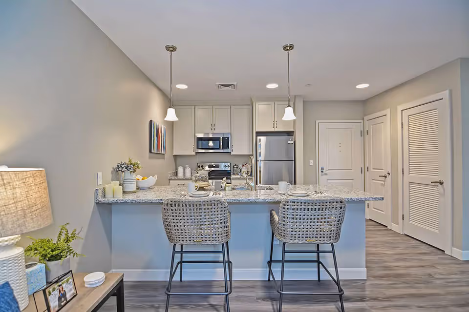 Modern kitchen area in a retirement community with a granite countertop island, two woven bar stools, stainless steel appliances including a refrigerator and microwave, pendant lights hanging above the island, and neutral-colored walls and flooring.